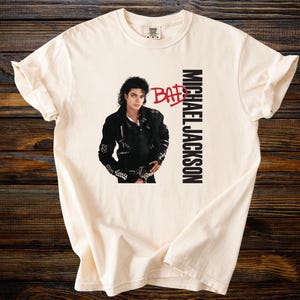 May include: Cream-colored t-shirt featuring a graphic of Michael Jackson in a black leather jacket. The word "BAD" is in red, and "MICHAEL JACKSON" is printed vertically in black. The shirt is on a dark wood surface.