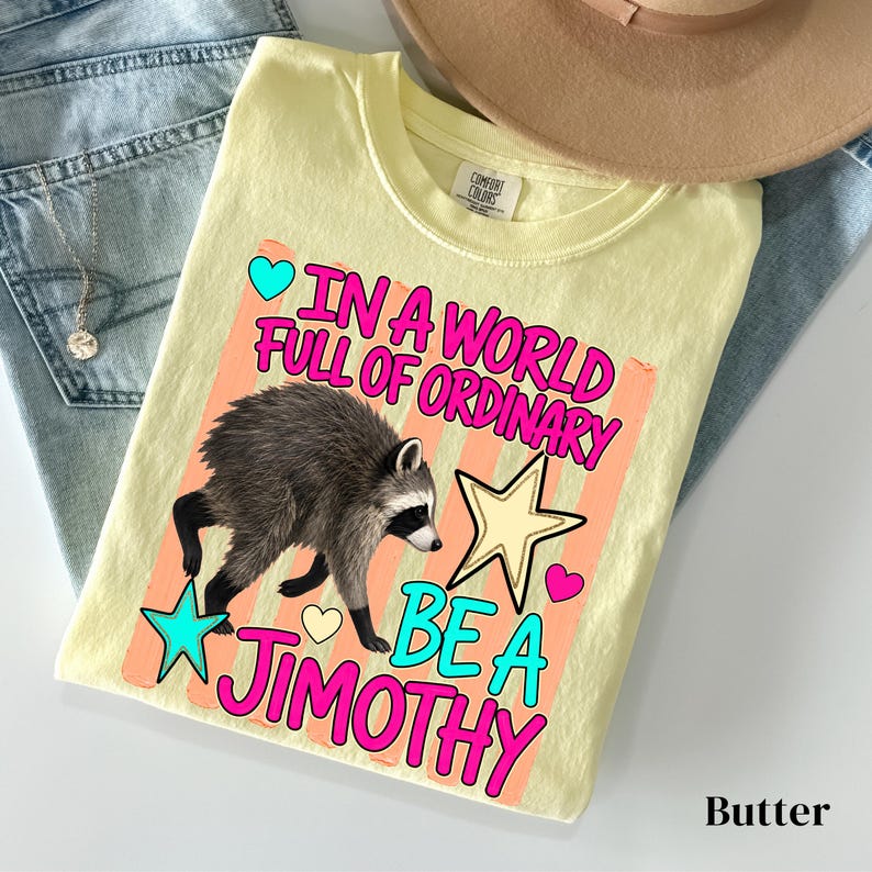 May include: A pale yellow t-shirt featuring a graphic of a raccoon and the text "IN A WORLD FULL OF ORDINARY BE A JIMOTHY" in bright pink and teal. The design includes a large yellow star, a small teal star, and two small hearts.