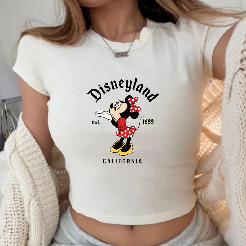Może przedstawiać: Kremowa koszulka z kr&oacute;tkim rękawem z grafiką Disneyland. Grafika zawiera Minnie Mouse w czerwonej sukience w kropki, czerwoną kokardę i napis "Disneyland est. 1955 CALIFORNIA."