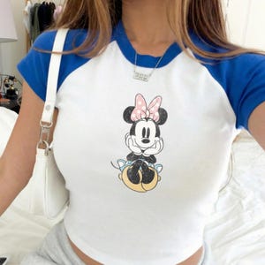 May include: White and blue raglan style t-shirt featuring a Minnie Mouse graphic. The shirt has a white body with blue sleeves and collar. The Minnie Mouse graphic shows Minnie Mouse with a pink bow and polka dots.