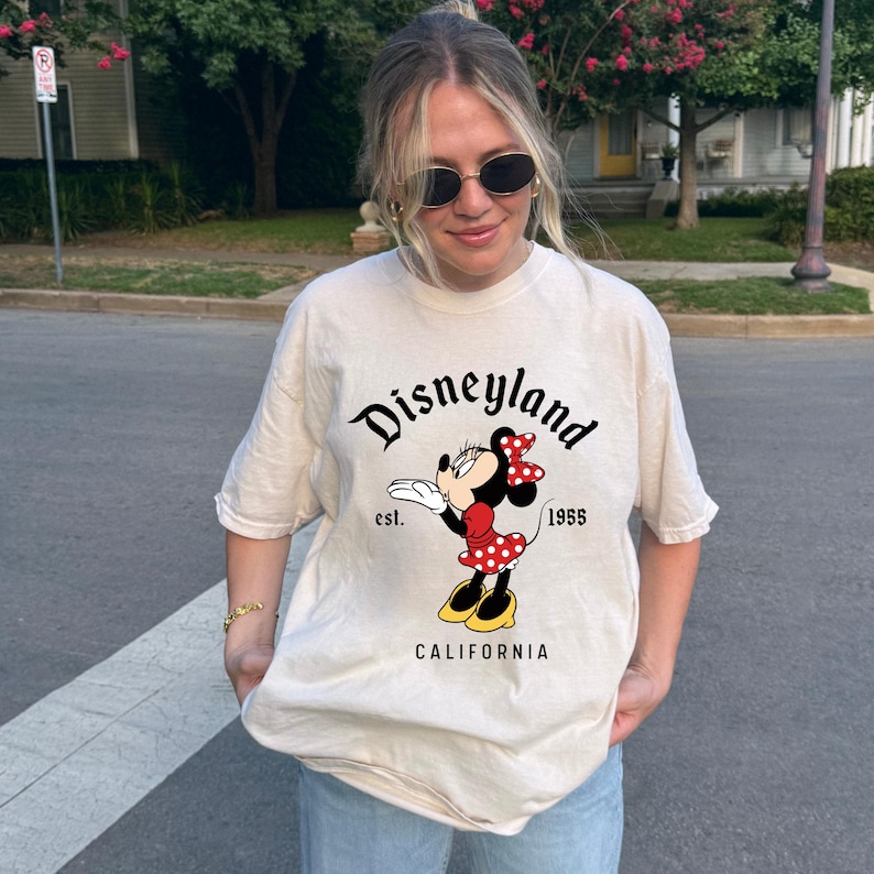 May include: Cream-colored t-shirt featuring the word "Disneyland" in black script above a Minnie Mouse graphic. Minnie is wearing a red dress with white polka dots and a matching bow. The shirt also includes the text "est. 1955 CALIFORNIA".