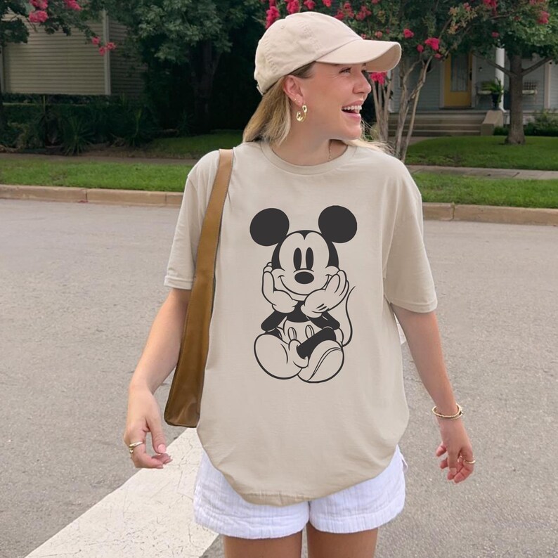 May include: Beige t-shirt featuring a black and white graphic of Mickey Mouse. The shirt has a relaxed fit and short sleeves. The person is wearing a beige baseball cap, gold earrings, and white shorts.