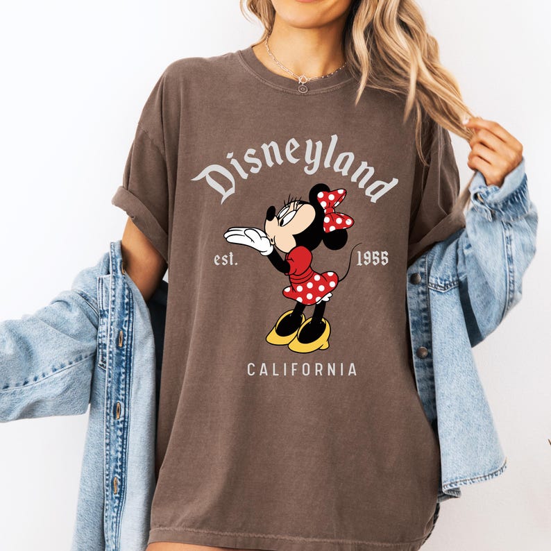 May include: Brown t-shirt featuring Minnie Mouse blowing a kiss. The design includes the text "Disneyland" above the character, with "est. 1955" and "CALIFORNIA" below. Minnie Mouse is wearing a red dress with white polka dots and a red bow.