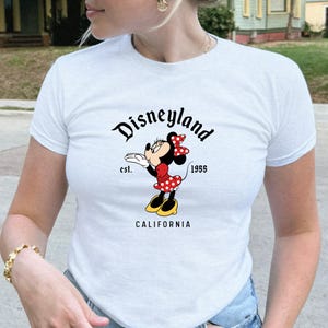 Może przedstawiać: Biała koszulka z grafiką Myszki Minnie. Projekt zawiera napis "Disneyland" nad kresk&oacute;wkową Myszką Minnie w czerwonej sukience w kropki, z napisem "est. 1955" i "CALIFORNIA" poniżej.