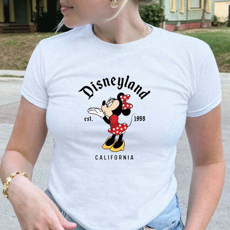 Może przedstawiać: Biała koszulka z grafiką Myszki Minnie. Projekt zawiera napis "Disneyland" nad kresk&oacute;wkową Myszką Minnie w czerwonej sukience w kropki, z napisem "est. 1955" i "CALIFORNIA" poniżej.