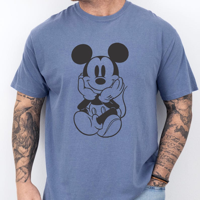 May include: A blue t-shirt featuring a black graphic of Mickey Mouse with his hands under his chin. The cartoon character is smiling. The shirt has short sleeves and a crew neck.
