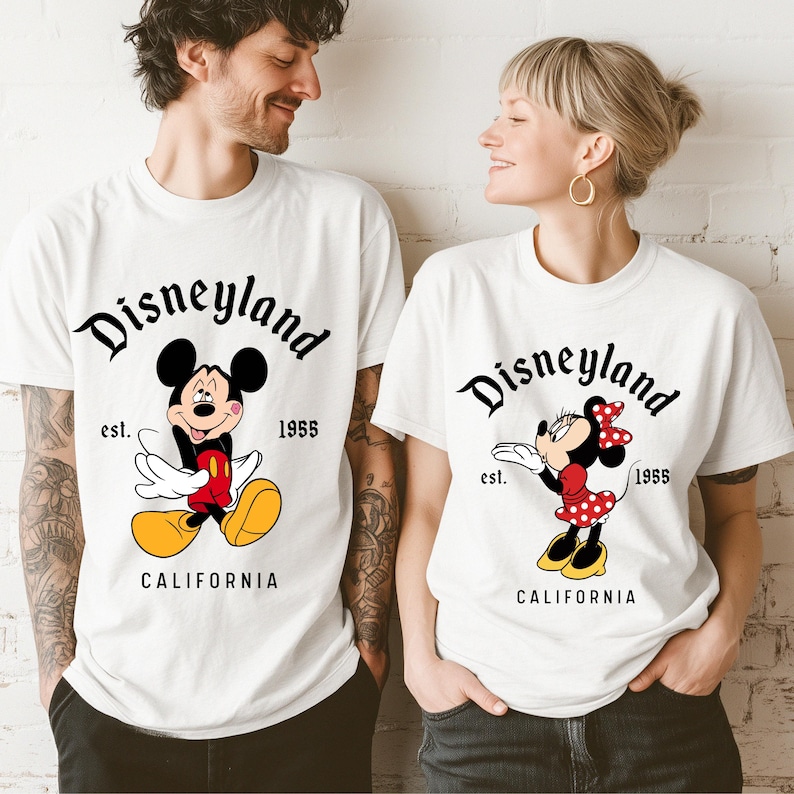 May include: White t-shirts with Disneyland graphics. One shirt features Mickey Mouse in red, yellow, and black, and the other features Minnie Mouse in a red polka-dot dress. Both shirts have the text "Disneyland" and "California".