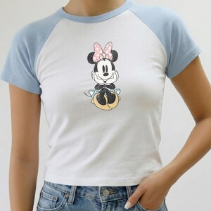 May include: White and light blue raglan t-shirt featuring a Minnie Mouse graphic. The graphic shows Minnie Mouse with a pink bow. The shirt has short sleeves and a crew neck. The model is wearing blue jeans.