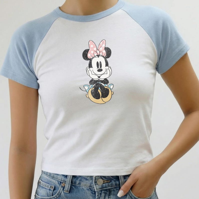 May include: White and light blue raglan t-shirt featuring a Minnie Mouse graphic. The graphic shows Minnie Mouse with a pink bow. The shirt has short sleeves and a crew neck. The model is wearing blue jeans.
