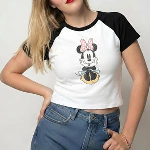 May include: White crop top with black sleeves featuring a Minnie Mouse graphic. The cartoon character is depicted with a pink bow and is sitting down. The shirt is paired with blue jeans.