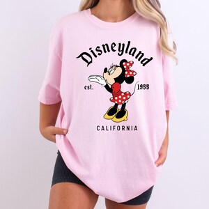 May include: A light pink t-shirt featuring a graphic of Minnie Mouse blowing a kiss. The design includes the text "Disneyland" in a script font, "est. 1955", and "CALIFORNIA". Minnie Mouse is wearing a red dress with white polka dots and a matching bow.
