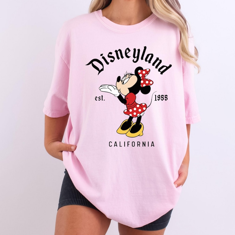 May include: A light pink t-shirt featuring a graphic of Minnie Mouse blowing a kiss. The design includes the text "Disneyland" in a script font, "est. 1955", and "CALIFORNIA". Minnie Mouse is wearing a red dress with white polka dots and a matching bow.