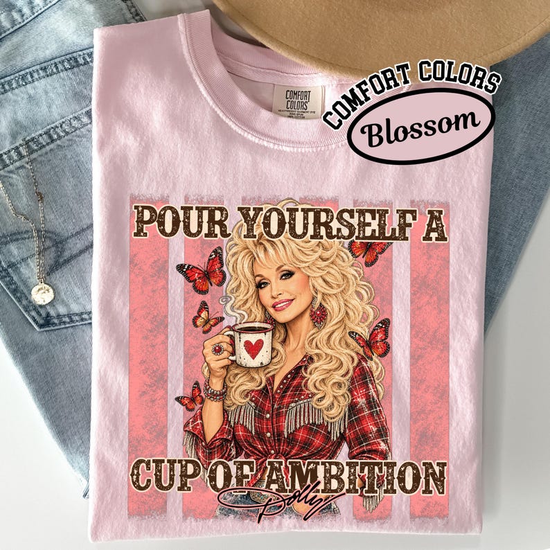 May include: A light pink t-shirt featuring a graphic of a blonde person with voluminous hair wearing a red plaid shirt, holding a white mug with a red heart. The text reads, "POUR YOURSELF A CUP OF AMBITION" with orange butterflies surrounding the figure.