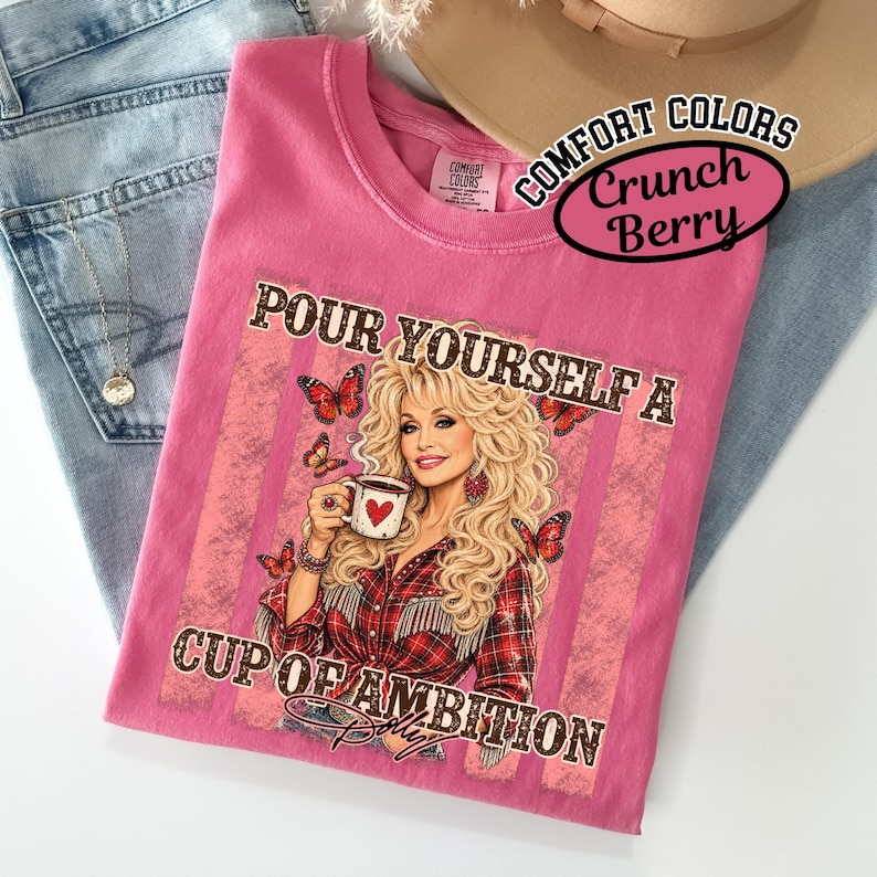 May include: A pink t-shirt featuring a graphic of a blonde person with long wavy hair wearing a red plaid shirt, holding a white mug with a red heart. The text reads "POUR YOURSELF A CUP OF AMBITION" with orange butterflies and pink stripes.