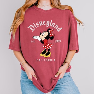 May include: A maroon t-shirt featuring a Minnie Mouse graphic. The design includes the word "Disneyland" in a script font, the year "1955", and the word "California". Minnie Mouse is wearing a red polka dot dress and bow.