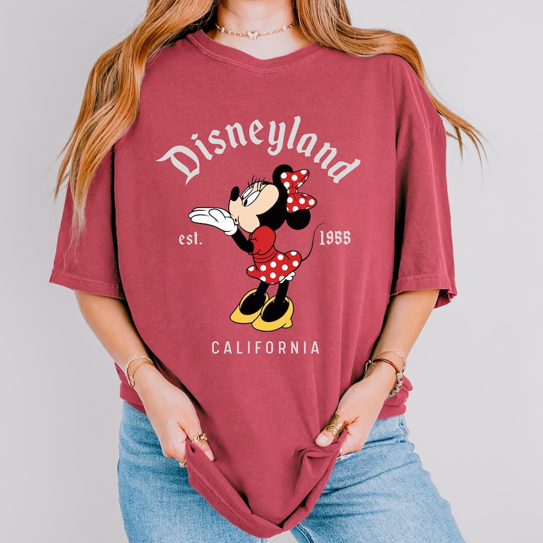 May include: A maroon t-shirt featuring a Minnie Mouse graphic. The design includes the word "Disneyland" in a script font, the year "1955", and the word "California". Minnie Mouse is wearing a red polka dot dress and bow.