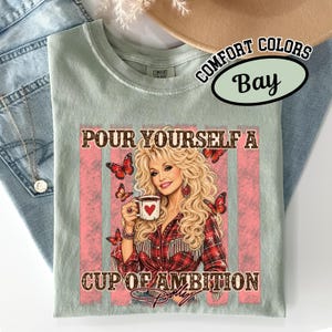 May include: A light green short-sleeve t-shirt featuring a graphic print of a blonde woman holding a coffee mug with a heart. The text reads "POUR YOURSELF A CUP OF AMBITION" with butterflies surrounding the figure. The shirt has a crew neckline.
