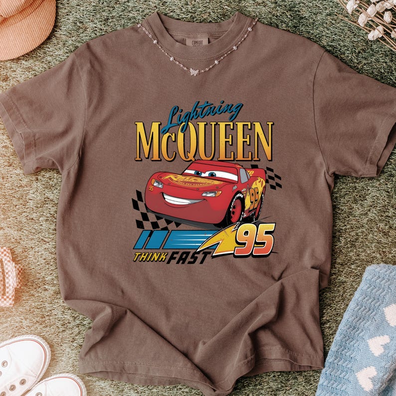 May include: Brown t-shirt featuring a graphic of Lightning McQueen from the movie Cars. The design includes the text "Lightning McQueen", the number "95", and the phrase "Think Fast".