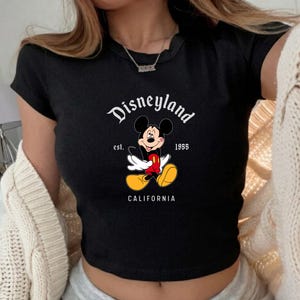Może przedstawiać: Czarny top z grafiką Myszki Miki i napisem "Disneyland" powyżej oraz "California" poniżej. Projekt zawiera napis "est. 1955". Koszulka ma kr&oacute;tkie rękawy i prążkowaną fakturę.