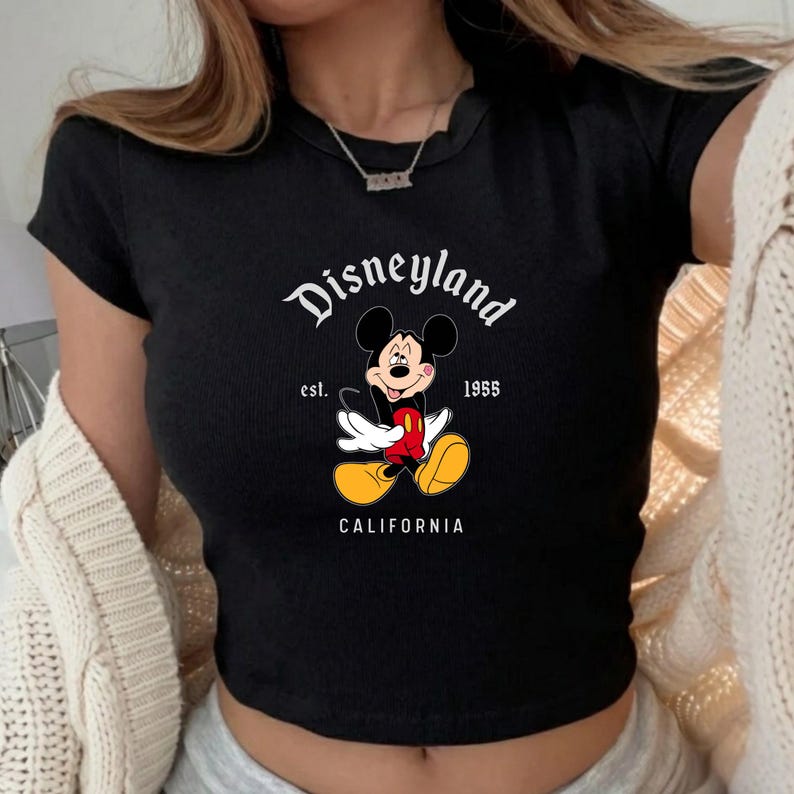 Może przedstawiać: Czarny top z grafiką Myszki Miki i napisem "Disneyland" powyżej oraz "California" poniżej. Projekt zawiera napis "est. 1955". Koszulka ma kr&oacute;tkie rękawy i prążkowaną fakturę.