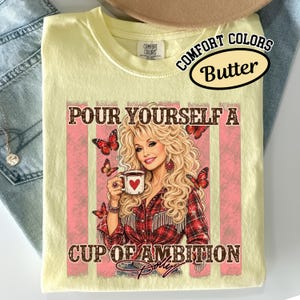 May include: A pale yellow t-shirt featuring a graphic of a blonde person with long wavy hair wearing a red plaid shirt. The person holds a white mug with a red heart. Text reads: POUR YOURSELF A CUP OF AMBITION. Red butterflies surround the figure.