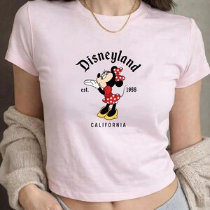 Może przedstawiać: Jasnor&oacute;żowa koszulka z grafiką Myszki Minnie w czerwonej sukience w grochy i kokardą. Tekst "Disneyland California est. 1955" jest wydrukowany nad i pod grafiką Myszki Minnie.