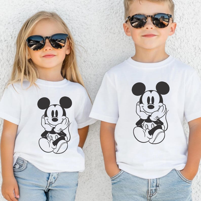 May include: Two children wearing white t-shirts with a black and white Mickey Mouse graphic. The graphic shows Mickey Mouse with his hands under his chin. Both children are wearing sunglasses and blue jeans.