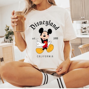May include: Cream-colored t-shirt featuring a classic Mickey Mouse graphic. The design includes the word "Disneyland" in a stylized font, along with "est. 1955" and "CALIFORNIA". The iconic cartoon character is depicted in a cheerful pose.