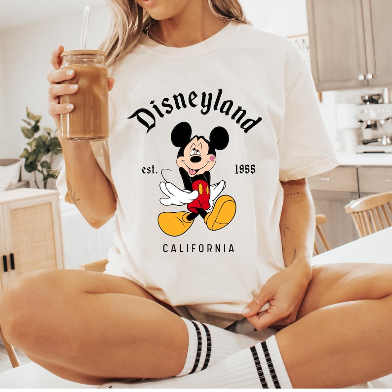 May include: Cream-colored t-shirt featuring a classic Mickey Mouse graphic. The design includes the word "Disneyland" in a stylized font, along with "est. 1955" and "CALIFORNIA". The iconic cartoon character is depicted in a cheerful pose.