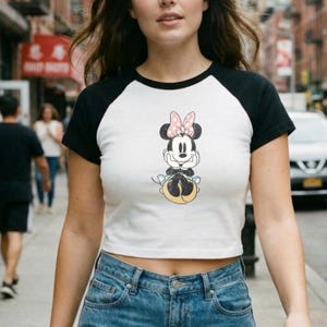May include: White crop top with black raglan sleeves featuring a Minnie Mouse graphic. The cartoon character is depicted with a pink bow and is seated. The top is paired with blue denim jeans.