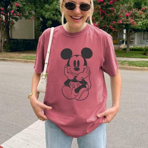 May include: A maroon t-shirt featuring a black graphic of Mickey Mouse. The cartoon character is depicted with his hands under his chin. The shirt is made of a soft material and is suitable for casual wear.