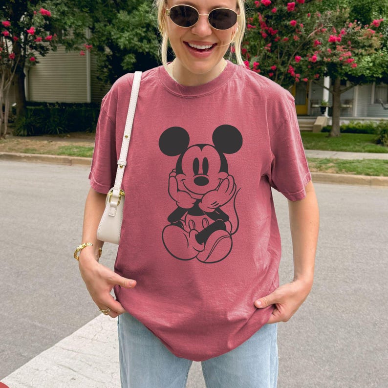 May include: A maroon t-shirt featuring a black graphic of Mickey Mouse. The cartoon character is depicted with his hands under his chin. The shirt is made of a soft material and is suitable for casual wear.