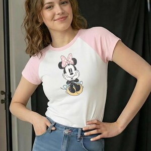 May include: White and pink raglan t-shirt featuring a Minnie Mouse graphic. The cartoon character is depicted with a pink bow and is resting her chin on her hands. The shirt is paired with blue jeans.