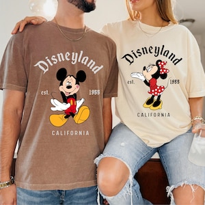 May include: Two t-shirts featuring the Disneyland logo and cartoon characters. One shirt is brown with Mickey Mouse, and the other is cream with Minnie Mouse. Both shirts have the text "Disneyland" and "California".