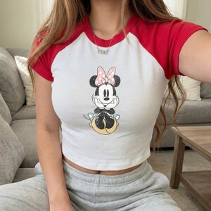 May include: White crop top with red short sleeves featuring a Minnie Mouse graphic. The cartoon character is depicted with a pink bow and is resting on a yellow and blue base. The top is a casual style.