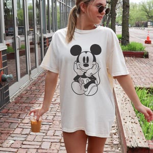 May include: Cream-colored t-shirt featuring a black and white graphic of Mickey Mouse. The oversized tee has short sleeves and a relaxed fit. The design shows Mickey Mouse with his hands under his chin, in a thoughtful pose.