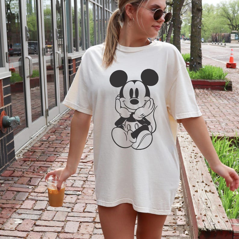 May include: Cream-colored t-shirt featuring a black and white graphic of Mickey Mouse. The oversized tee has short sleeves and a relaxed fit. The design shows Mickey Mouse with his hands under his chin, in a thoughtful pose.