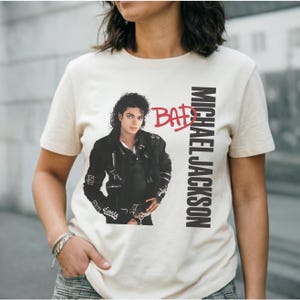 May include: Cream-colored t-shirt featuring a portrait of Michael Jackson in a black jacket. The word "BAD" is in red above the image, with "MICHAEL JACKSON" printed vertically in black on the right side.