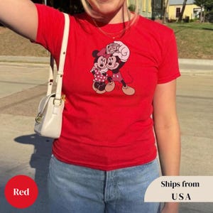 Pu&ograve; includere: Una maglietta rossa con un'immagine a cartoni animati di Topolino e Minnie. Minnie indossa un vestito a pois e Topolino tiene una macchina fotografica. La maglietta &egrave; indossata da una persona.