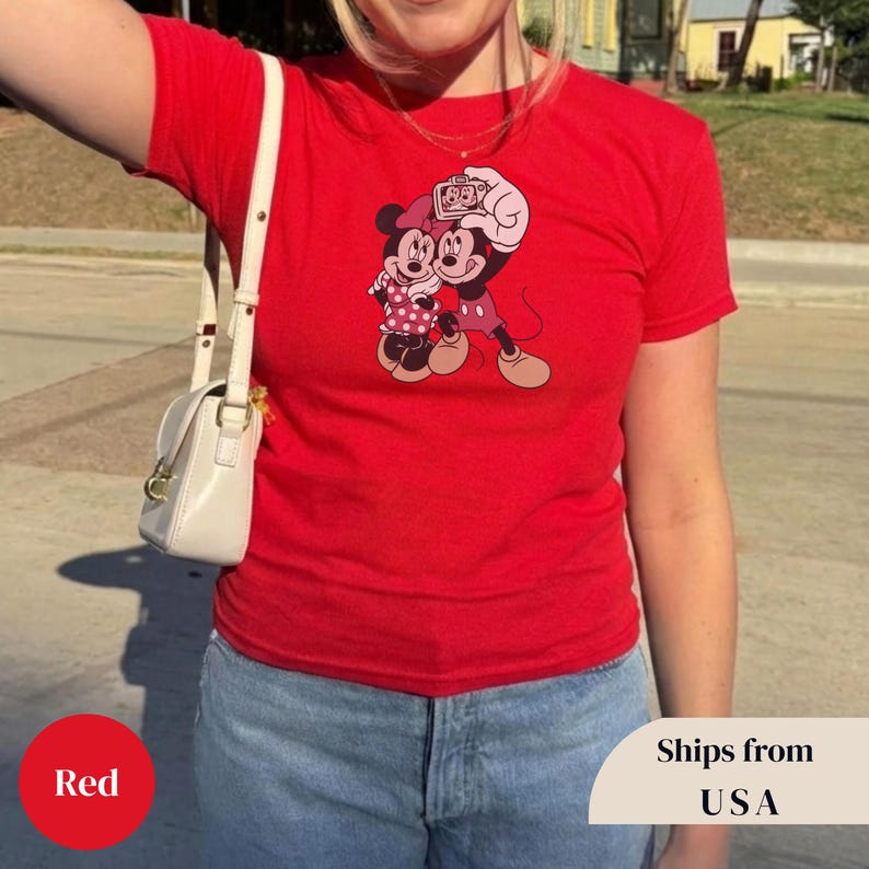 Pu&ograve; includere: Una maglietta rossa con un'immagine a cartoni animati di Topolino e Minnie. Minnie indossa un vestito a pois e Topolino tiene una macchina fotografica. La maglietta &egrave; indossata da una persona.