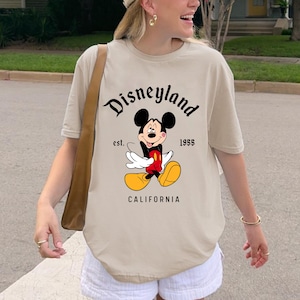 May include: Beige t-shirt featuring a graphic of Mickey Mouse with the text "Disneyland" above and "est. 1955 CALIFORNIA" below. The shirt has short sleeves and a relaxed fit.