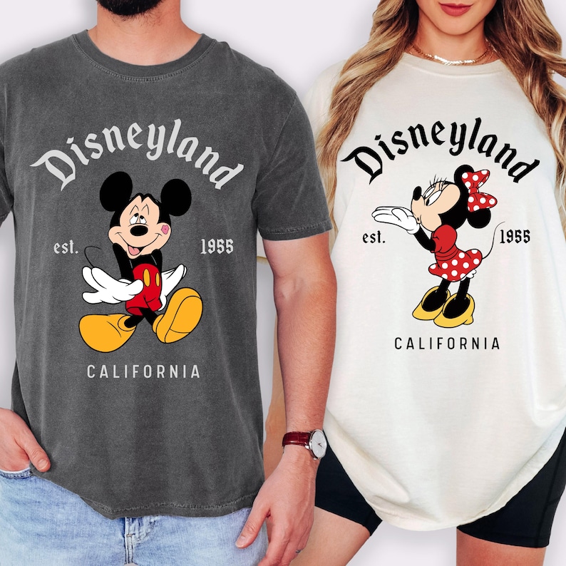 May include: Two t-shirts featuring the Disneyland logo and cartoon characters. One is dark gray with Mickey Mouse, the other is off-white with Minnie Mouse. Both shirts have the text "Disneyland" above the character, "est. 1955" and "CALIFORNIA" below.