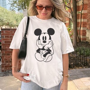May include: White t-shirt featuring a black and white graphic of Mickey Mouse. The cartoon character is seated with his hands under his chin. The shirt is a casual style, suitable for everyday wear.