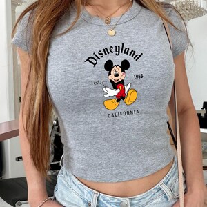 Może przedstawiać: Szara, kr&oacute;tka koszulka z nadrukiem Myszki Miki i napisem "Disneyland California 1955". Koszulka ma prążkowaną fakturę i okrągły dekolt.