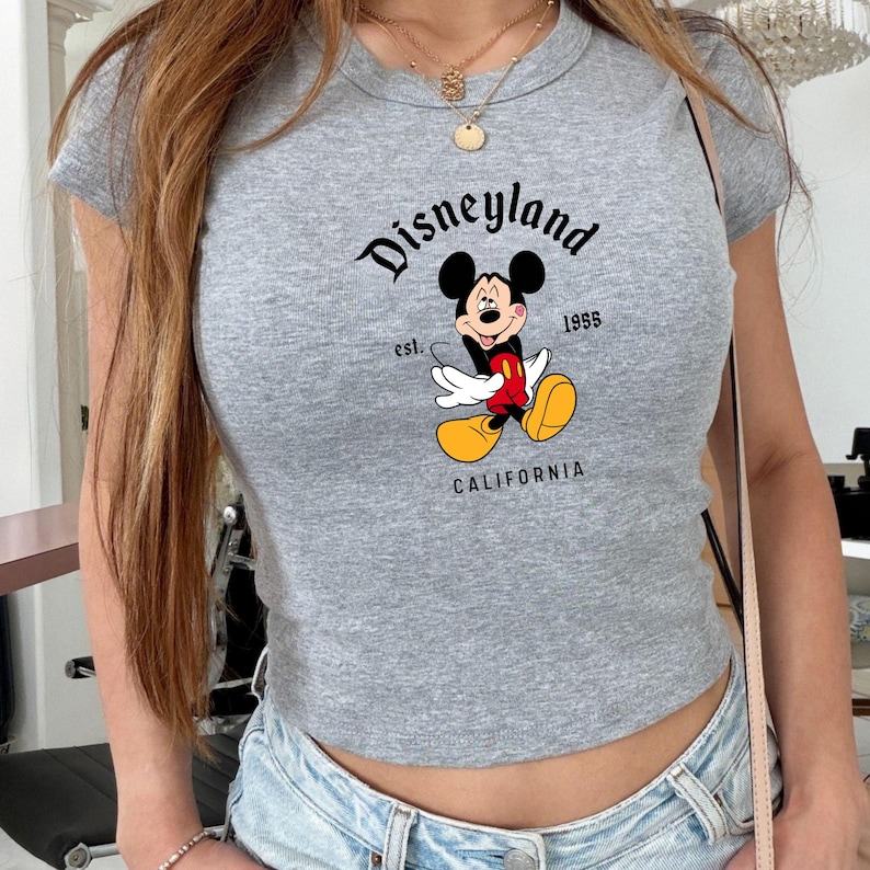 Może przedstawiać: Szara, kr&oacute;tka koszulka z nadrukiem Myszki Miki i napisem "Disneyland California 1955". Koszulka ma prążkowaną fakturę i okrągły dekolt.