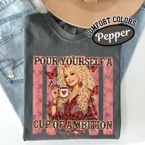 May include: A charcoal grey t-shirt featuring a graphic of a blonde woman holding a coffee mug with a heart. The design includes red butterflies, a plaid shirt, and the text "POUR YOURSELF A CUP OF AMBITION" in a brown, distressed font style.
