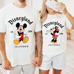 May include: Off-white t-shirts featuring Mickey and Minnie Mouse designs. The shirts have the text "Disneyland" above the characters, with "est. 1955" and "CALIFORNIA" below. Mickey is in red shorts and yellow shoes. Minnie wears a red polka dot dress.
