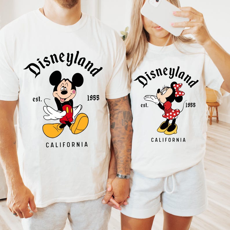 May include: Off-white t-shirts featuring Mickey and Minnie Mouse designs. The shirts have the text "Disneyland" above the characters, with "est. 1955" and "CALIFORNIA" below. Mickey is in red shorts and yellow shoes. Minnie wears a red polka dot dress.