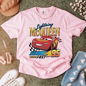 May include: A light pink t-shirt featuring a graphic of Lightning McQueen from the movie Cars. The design includes the text "Lightning McQueen", "Think Fast", and the number "95". The shirt has a crew neck and short sleeves.