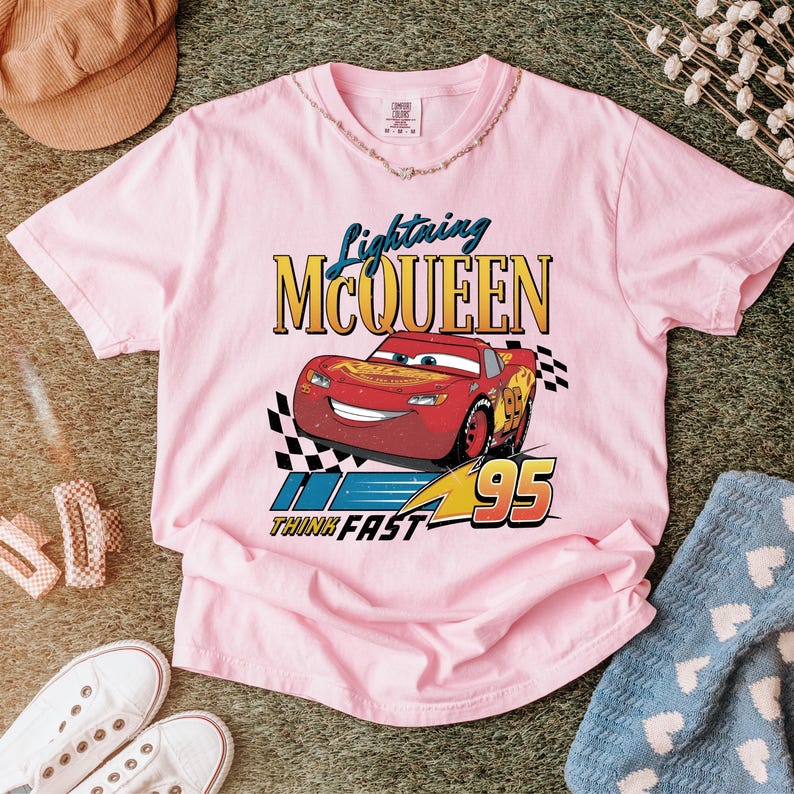 May include: A light pink t-shirt featuring a graphic of Lightning McQueen from the movie Cars. The design includes the text "Lightning McQueen", "Think Fast", and the number "95". The shirt has a crew neck and short sleeves.
